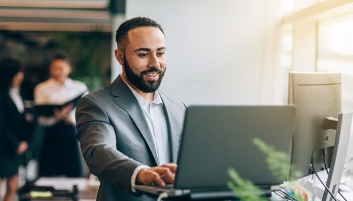 Man standing smiling while updating laptop in bright business office wearing suit